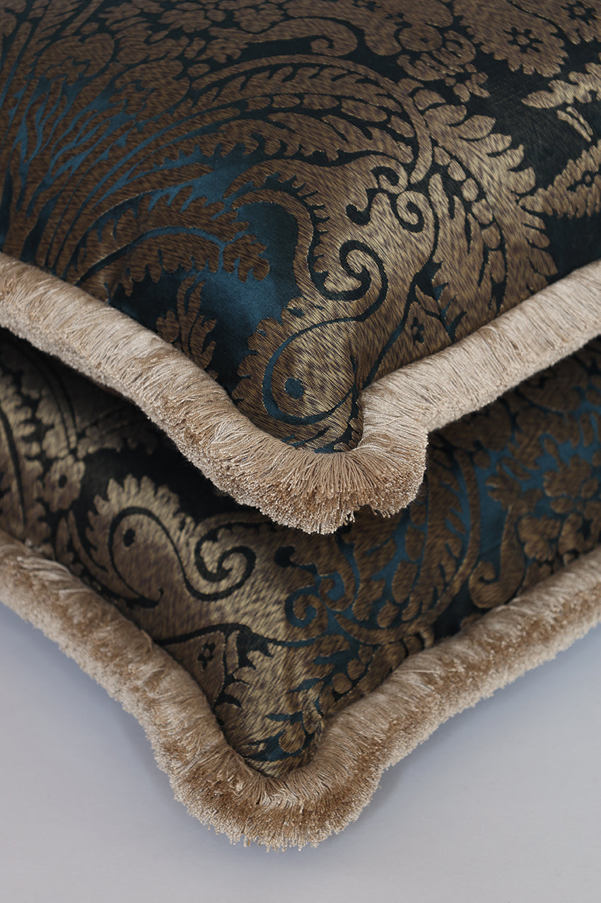 Close-up of a decorative throw pillow featuring a dark damask pattern with teal-blue undertones and a beige fringed border.