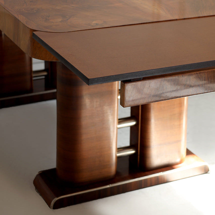 A close-up of a polished wooden dining table with a rich brown grain, featuring a drop-leaf design and a dark apron, set against a light neutral background.