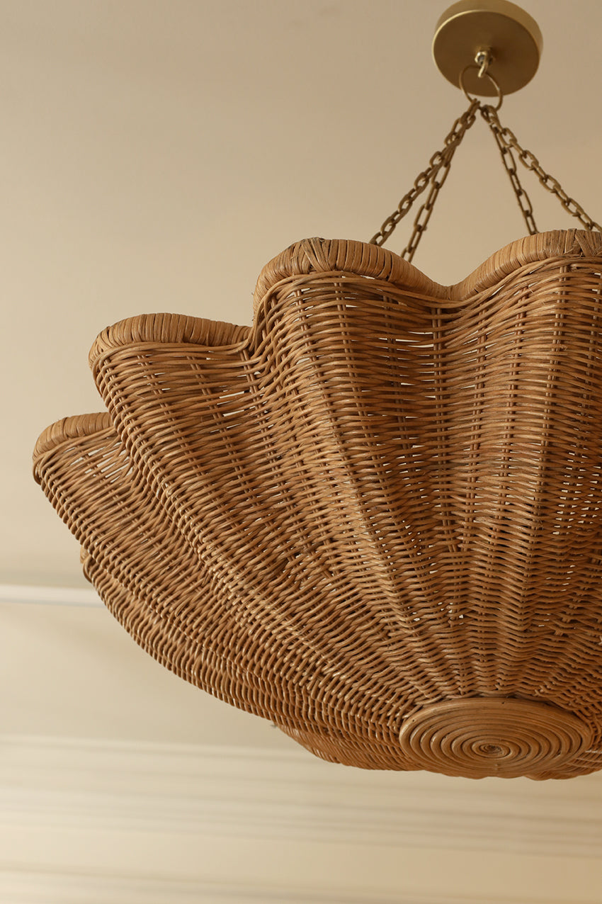 A large woven wicker chandelier pendant light in a natural tan color, suspended from a brass ceiling canopy with chains against a cream-colored wall.