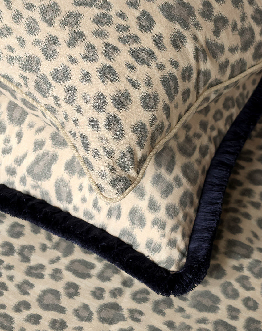 Close-up of a leopard-print throw pillow with a dark navy blue fringed edge, resting on a matching leopard-print fabric surface.