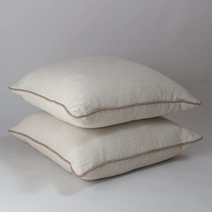 Two square off-white linen throw pillows stacked on top of each other, featuring a contrasting light brown piped edge, set against a neutral gray studio background.