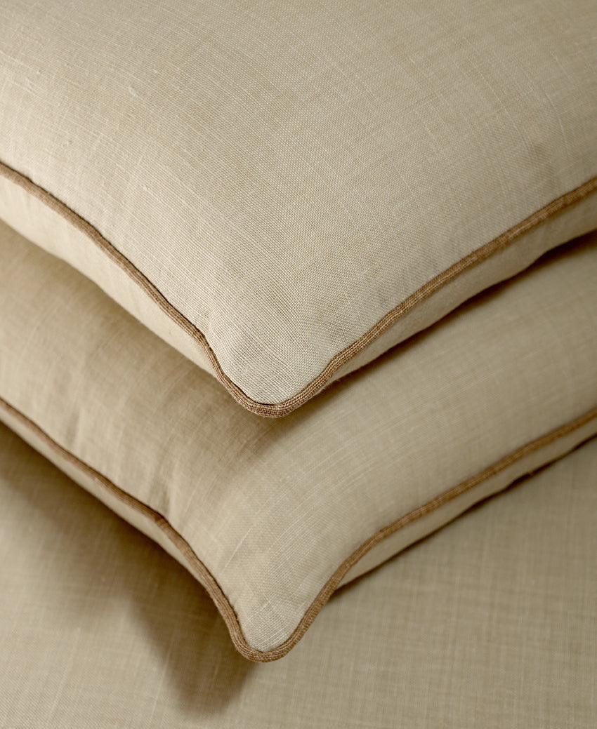 Two stacked beige linen pillows with contrasting brown piped edges, shown on a matching textured beige fabric background.