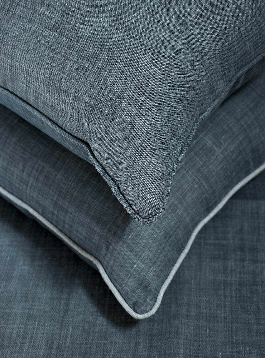 A close-up of a textured slate blue throw pillow resting on a matching blue fabric surface, showing a thin white piped edge.