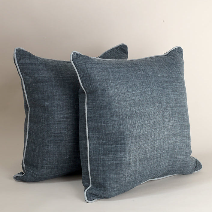 Two square throw pillows in a textured slate blue fabric with light piping, arranged against a neutral beige background.