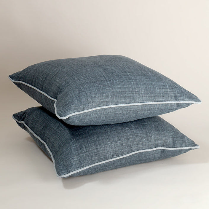 Two square throw pillows in a textured slate blue fabric with white piping, stacked on a light beige background.
