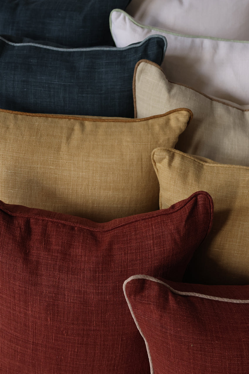 A close-up arrangement of textured throw pillows in multiple colors, including deep burgundy, mustard yellow, cream, navy, and light beige, with some showing white piping.