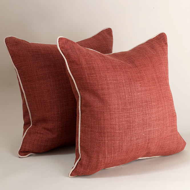 Two square throw pillows in a textured rust red fabric with a thin white piped edge, displayed against a neutral light beige background.