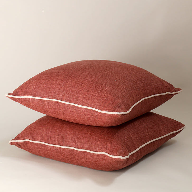 Two square throw pillows in a textured rust red fabric, stacked on top of each other with a thin white piping trim, set against a plain light beige background.