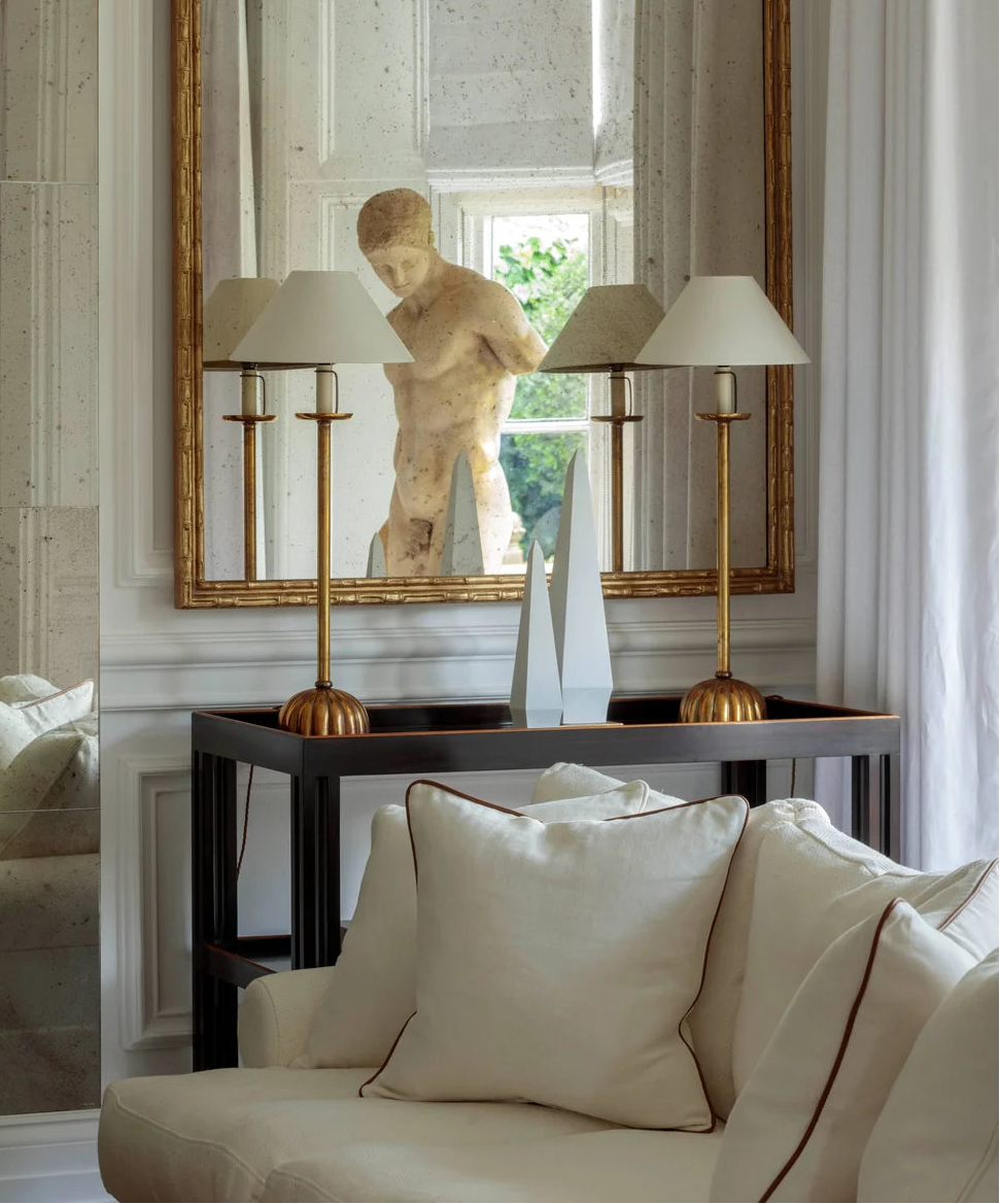 A large ornate gold-framed mirror hangs on a white paneled wall above a dark console table. The mirror reflects a classical-style stone bust sculpture, flanked by two brass table lamps with white drum shades.