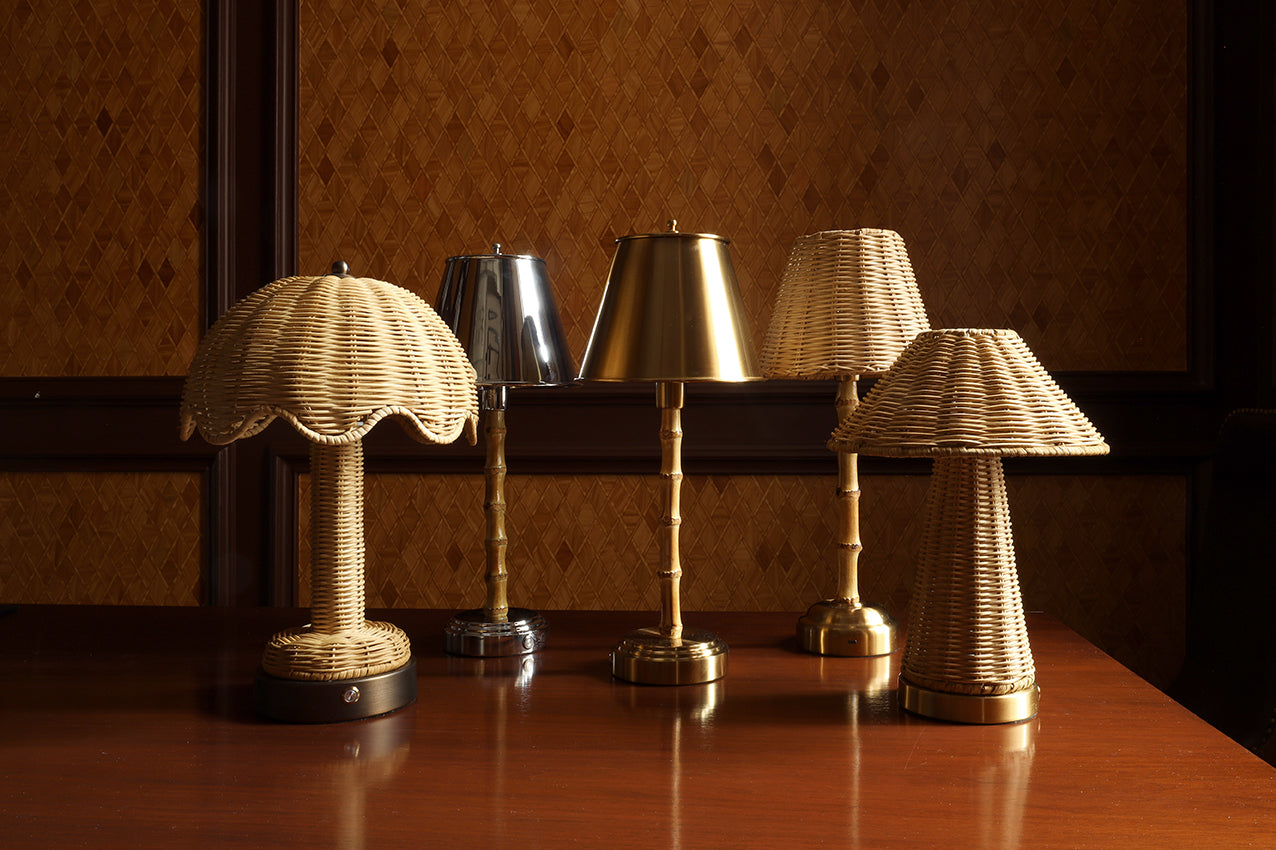 Four decorative table lamps with woven wicker shades and metallic brass bases arranged on a polished wooden table against a patterned brown wall.