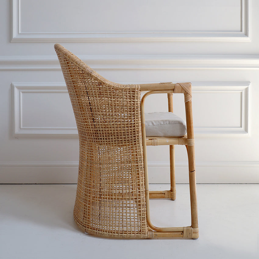 Woven rattan chair with a white cushion on a light-colored floor against a white paneled wall.