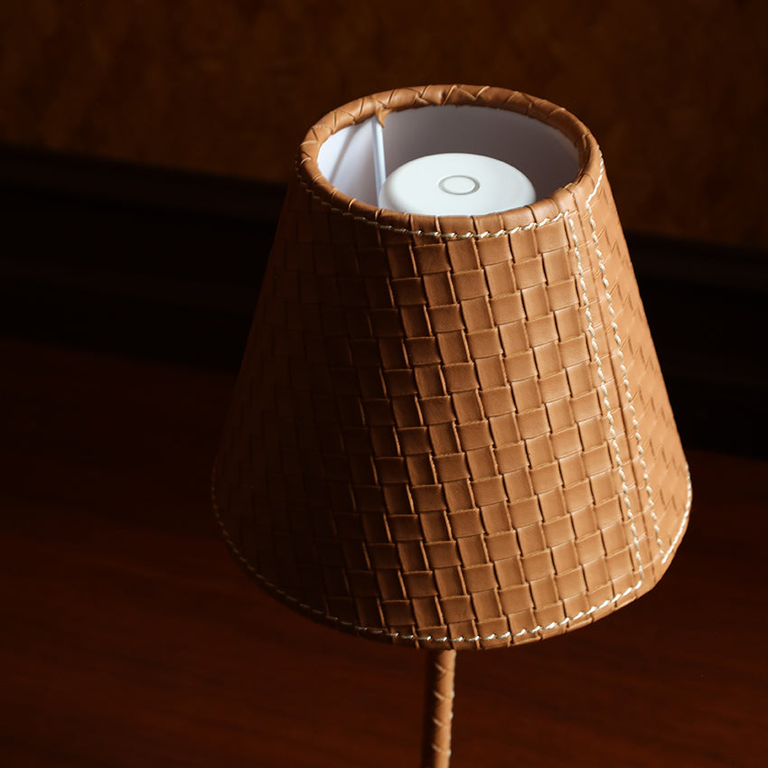 A woven brown lampshade with white contrast stitching, shown on a table lamp against a warm brown background.