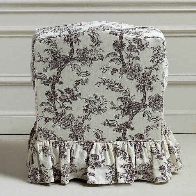 A ruffled fabric chair slipcover with a classic toile floral and bird pattern in dark gray on an off-white background, shown on a chair against a white paneled wall.