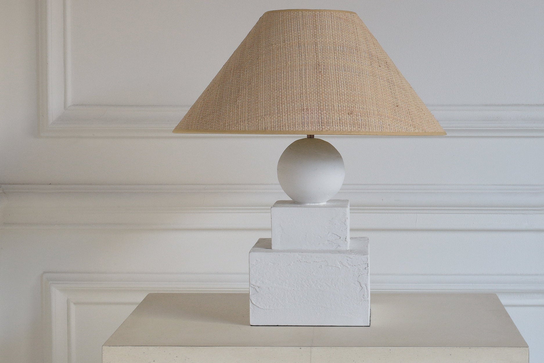 Table lamp with a beige shade on a white pedestal against a white paneled wall.