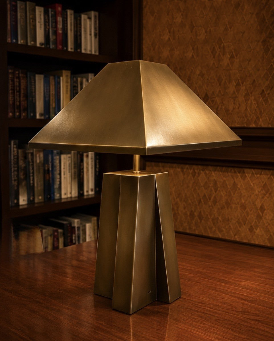 A modern table lamp with a geometric, faceted metallic bronze base and a matching wide, pyramid-shaped shade, set on a wooden table in a warm, book-filled room.