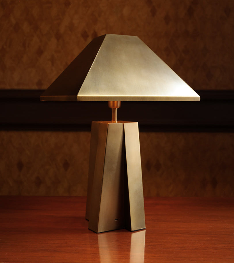 A modern table lamp with a geometric, angular metallic base and a matching triangular shade, shown on a wooden surface against a textured brown wall.