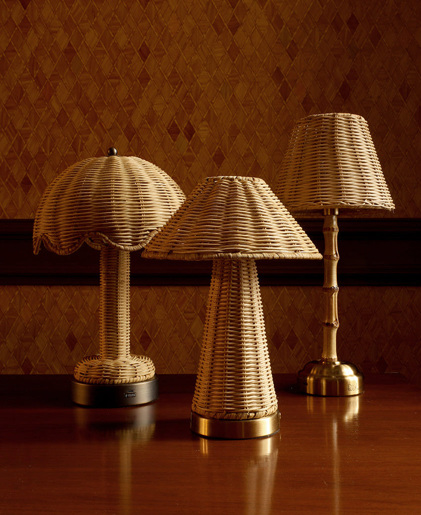 Three woven rattan table lamps with gold-finished bases and shades, arranged on a polished wooden table against a patterned brown wallpaper background.