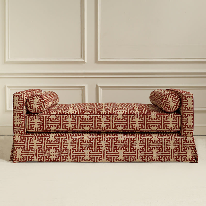 An upholstered bench with rolled arms and a skirted base, covered in a red and beige geometric patterned fabric, set against a cream paneled wall.