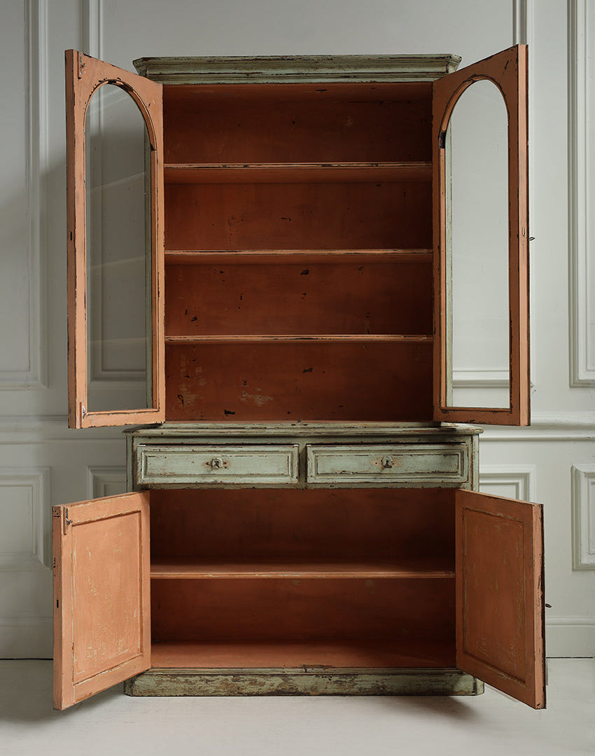 A tall vintage wooden hutch or cabinet with distressed paint, shown with all doors open to reveal multiple shelves and two upper cabinet doors with arched mirror insets. The cabinet has a warm brown wood interior and exterior, with a light weathered greenish-gray painted top and drawer section.