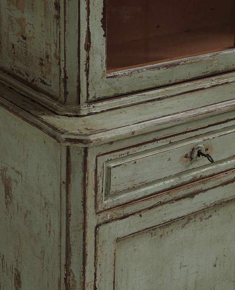 Close-up of a distressed vintage cabinet in a muted sage green paint with peeling layers showing the wood underneath, featuring a glass upper section and a single drawer with a small metal pull.