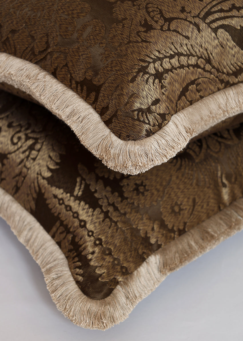 Close-up of a decorative throw pillow with a brown and beige damask pattern and a fringed edge, folded over on a light gray background.