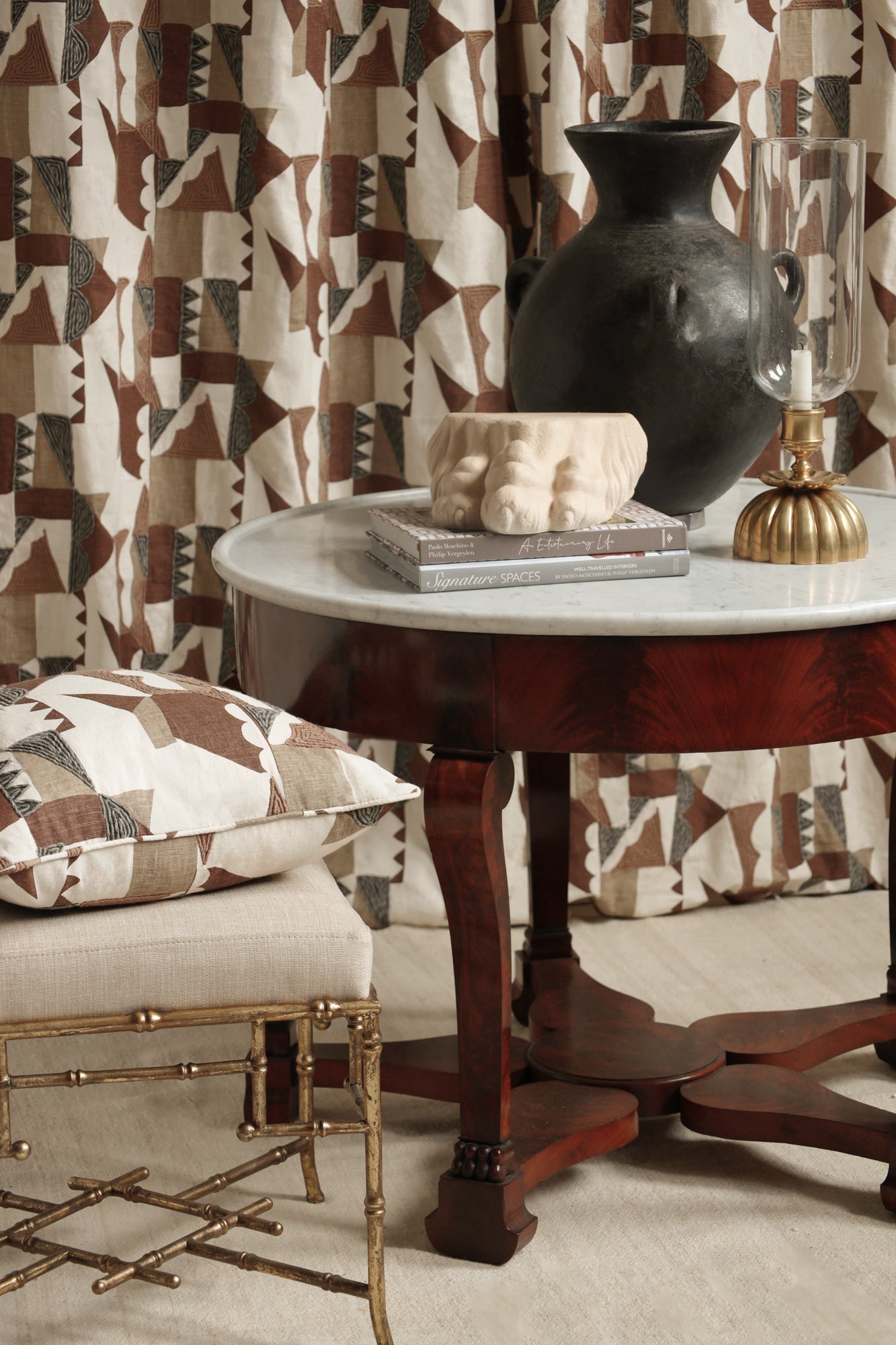 Decorative room setting with a round table, vase, and decorative items against patterned curtains.