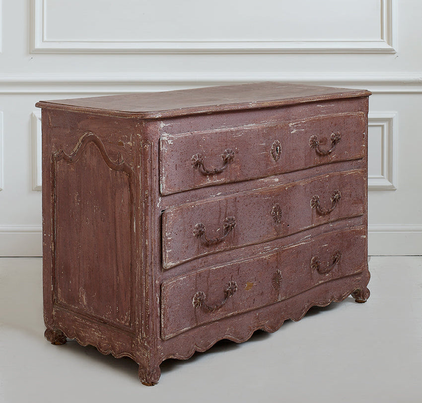 An antique wooden chest of drawers with a distressed reddish-brown finish, featuring three drawers with ornate metal handles and a scalloped front panel, set against a white paneled wall.