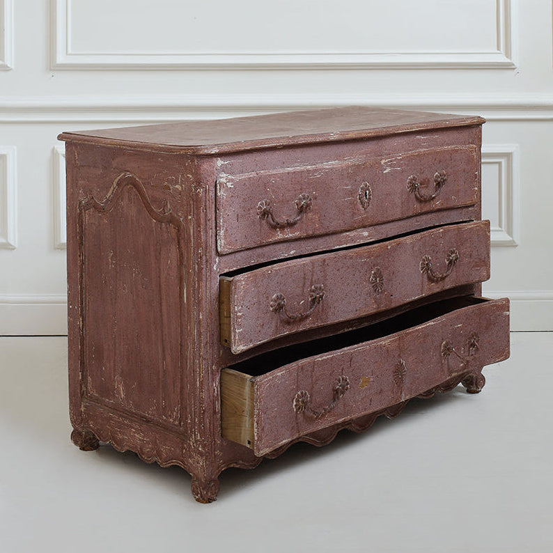 A distressed reddish-brown wooden chest of drawers with three drawers, one pulled open, set against a white paneled wall.