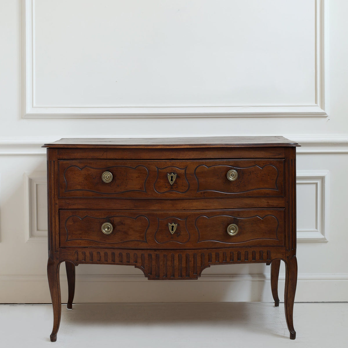 A vintage wooden chest of drawers with two drawers, ornate carved fronts, brass knobs, and curved cabriole legs, set against a white paneled wall.