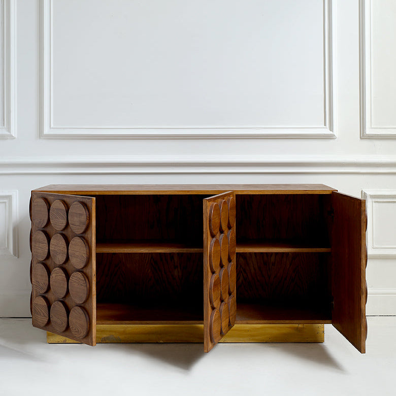 A mid-century modern wooden sideboard cabinet with two doors swung open, showcasing circular cutout details on the front panels and shelves inside, set against a white paneled wall.
