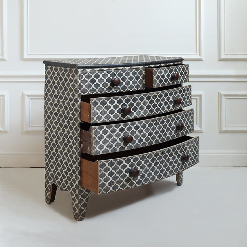A four-drawer chest of drawers with a black and white geometric trellis pattern and dark brown wooden drawer pulls, set against a white paneled wall.