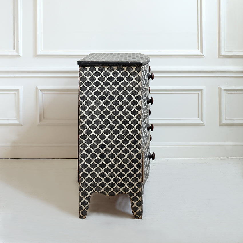 A small three-drawer chest or nightstand with a black and white geometric trellis pattern, featuring a dark top and dark drawer pulls, set against a white paneled wall.