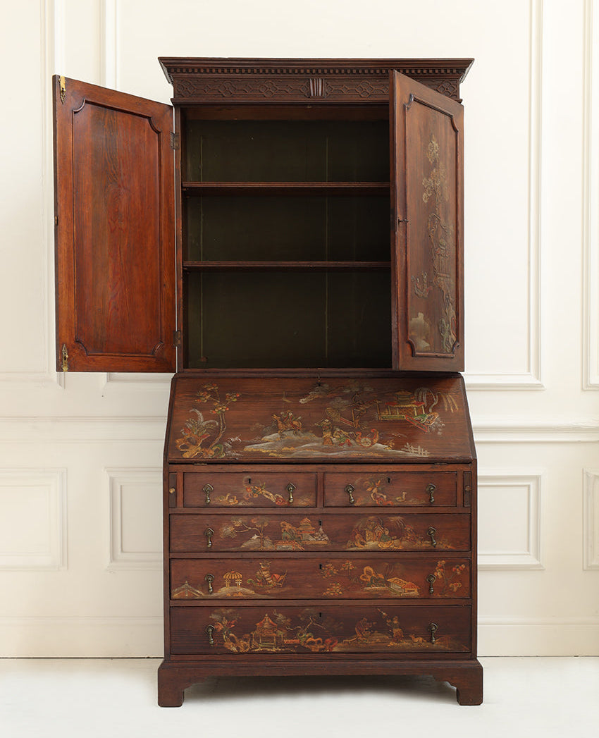 A dark reddish-brown wooden secretary desk cabinet with open upper doors and hand-painted scenic panels on the drawer fronts and writing surface, set against a white paneled wall.