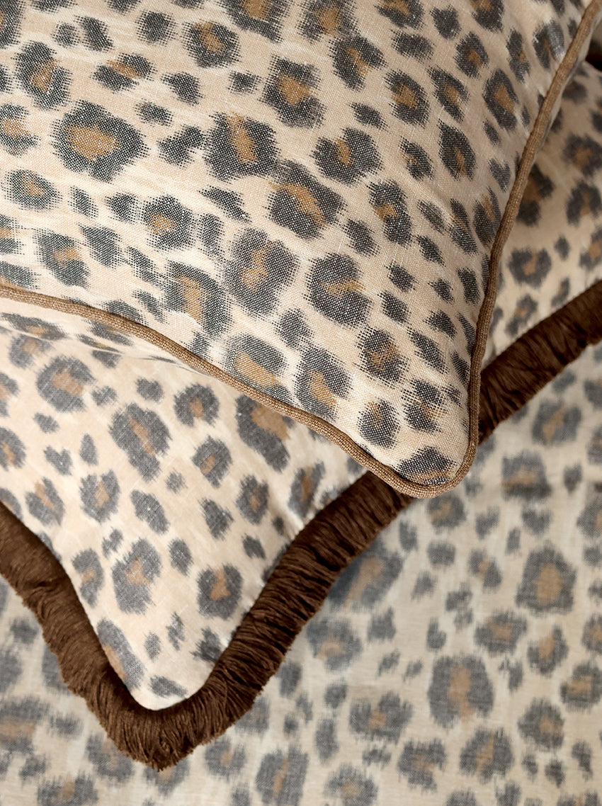 Close-up of leopard-print bedding with a cream base, brown and gray spots, and a dark brown fringed edge.