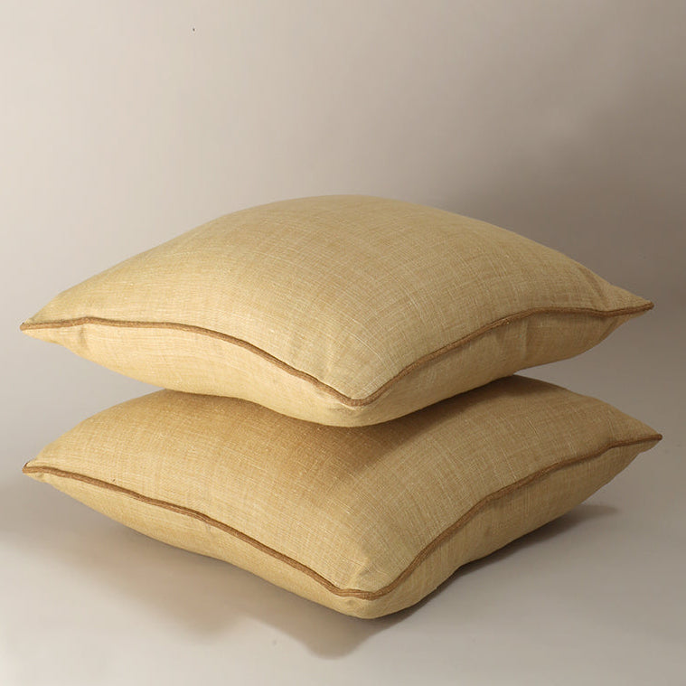 Two square beige throw pillows stacked on top of each other, made from a textured linen-like fabric with a thin brown piping edge, photographed against a neutral light gray studio background.