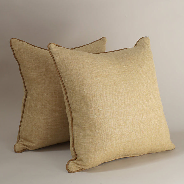 Two square decorative throw pillows in a warm beige linen-like fabric with brown piping, photographed on a plain light gray studio background.