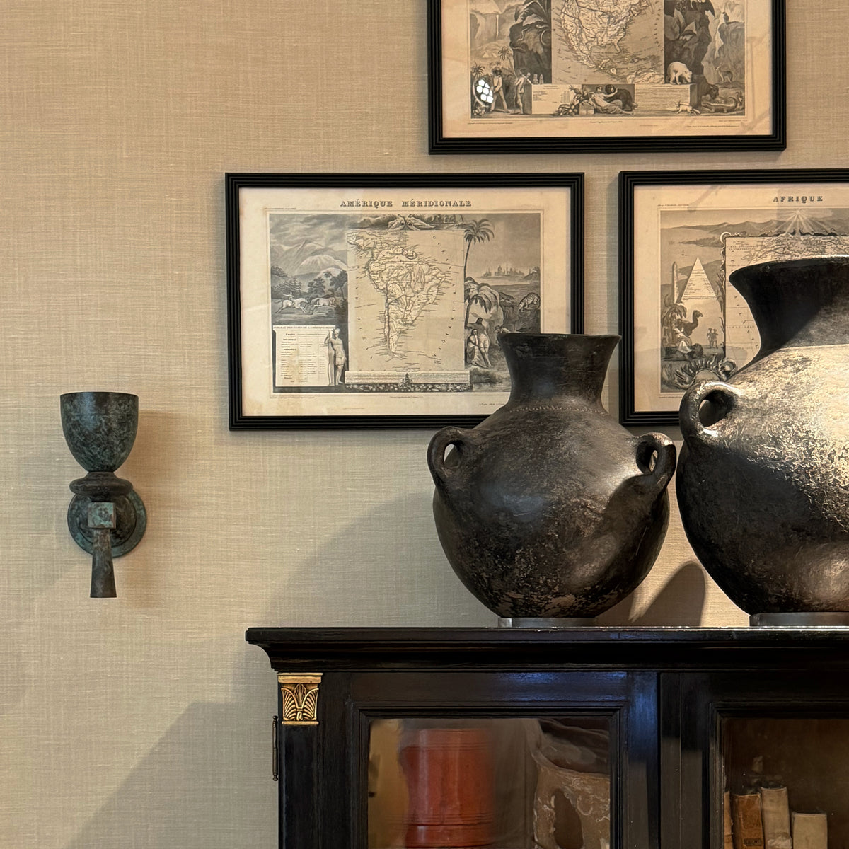 A styled interior scene showing framed vintage world maps on a textured beige wall above a dark wooden cabinet, with two large dark ceramic amphorae vases in the foreground and a bronze wall sconce on the left.