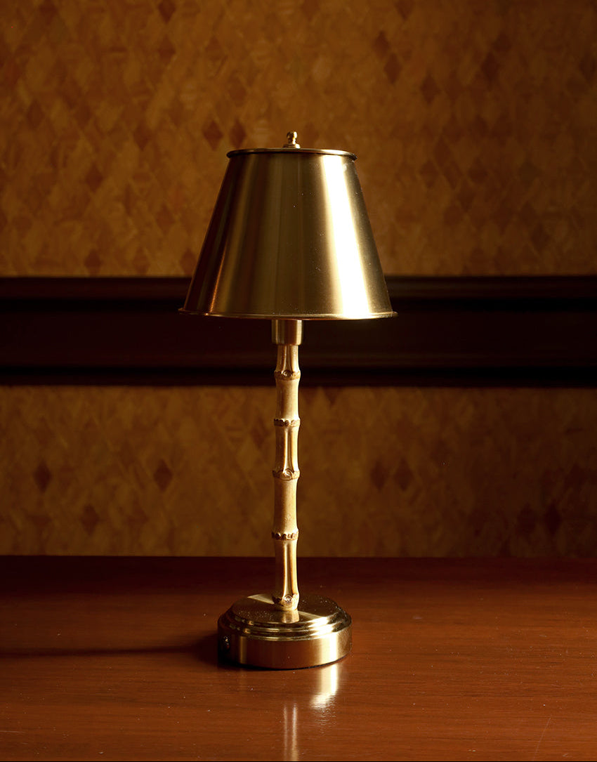 A brass table lamp with a conical shade and a bamboo-style jointed stem, standing on a polished wooden table against a patterned brown wallpaper background.