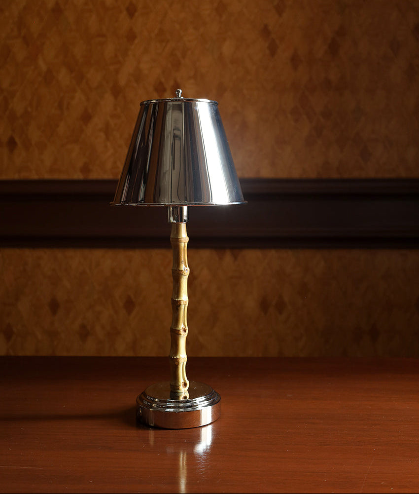 A small table lamp with a polished silver metal shade and base, and a twisted gold-toned stem, standing on a wooden surface against a textured brown wall.