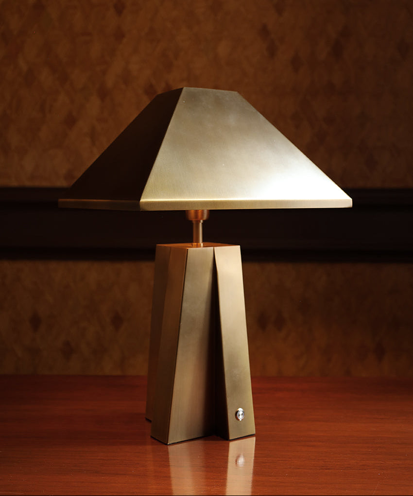 A modern table lamp with a geometric, faceted metallic base and a matching tapered shade, shown lit on a wooden surface against a textured brown wall.