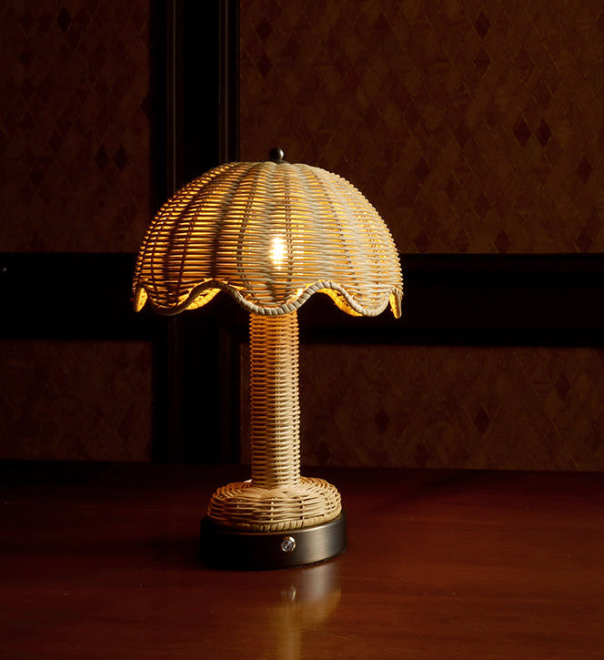 A small table lamp with a woven rattan or wicker shade and matching stem, illuminated with a warm yellow glow, set on a dark wooden surface against a dark patterned wall.