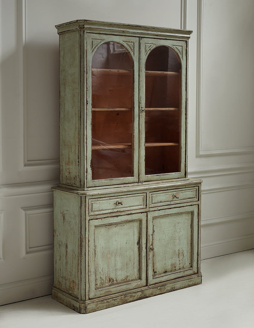 A distressed vintage-style hutch cabinet with a weathered sage green and beige paint finish, featuring two upper glass-paneled doors, two middle drawers, and two lower doors, set against a white paneled wall.