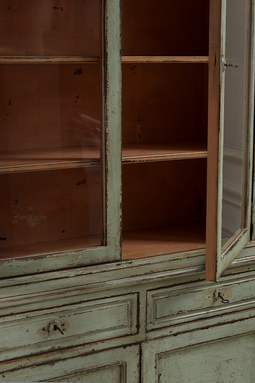 Close-up of a distressed vintage cabinet with a light sage green painted exterior and warm brown wood interior, featuring glass doors and open shelves.