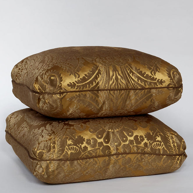 Two square decorative cushions stacked on top of each other, upholstered in a rich gold brocade fabric with an ornate floral and scroll pattern, photographed on a seamless light gray studio background.