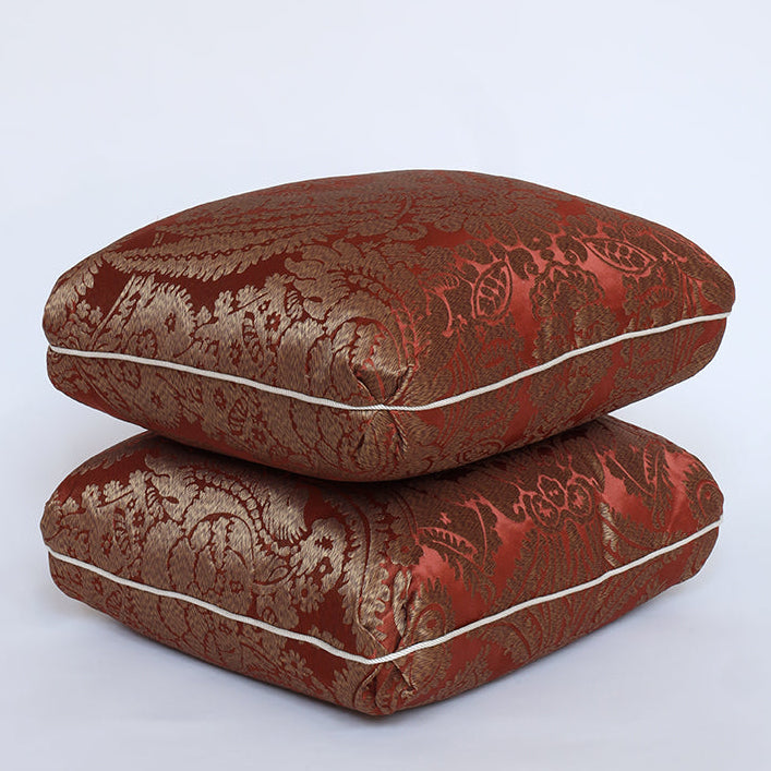 Two square decorative cushions stacked on a light gray background. The cushions feature a rich, metallic brocade pattern in deep red and gold, with a thin white piping detail along the edges.