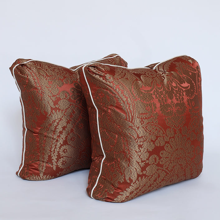 Two square decorative throw pillows in a rich red and gold brocade pattern with white piping, set against a plain light gray background.