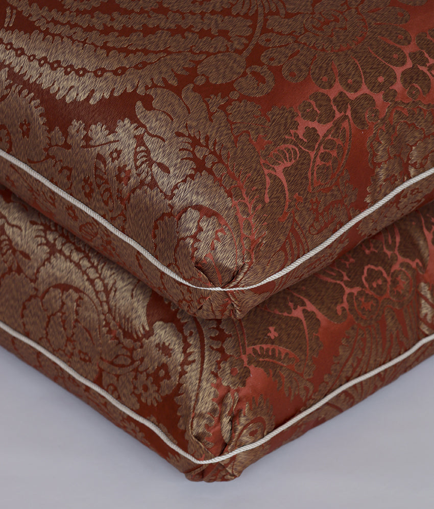 A close-up of a folded decorative pillow or cushion showing a rich damask pattern in rust red and gold, finished with white piping, set against a neutral light gray background.